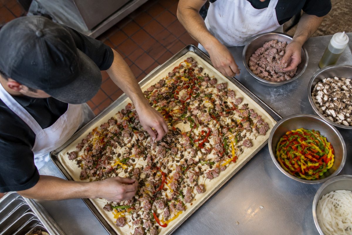 Kitchen crew assembling fresh toppings across a full-sheet banquet pizza pan